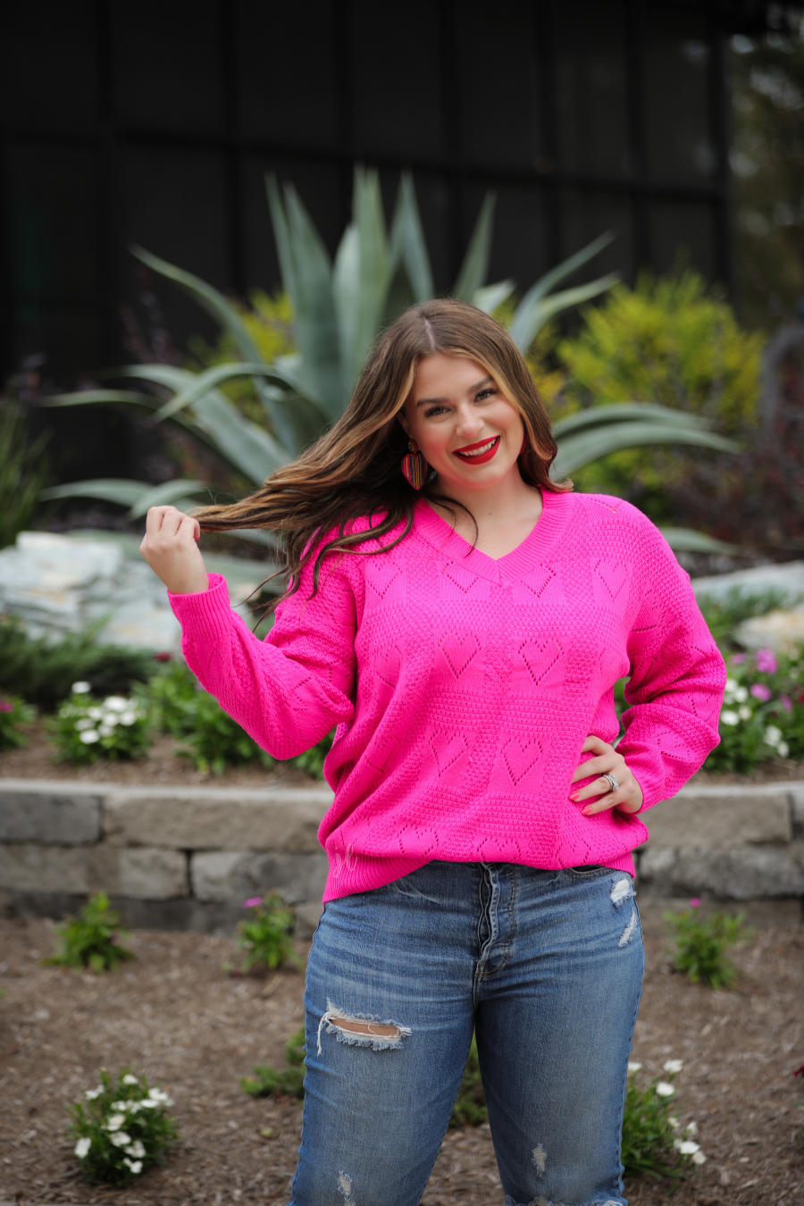 a pink long-sleeve open-knit sweater with a cutout heart pattern and ribbed detailing on the V-neckline, cuffs, and hemline