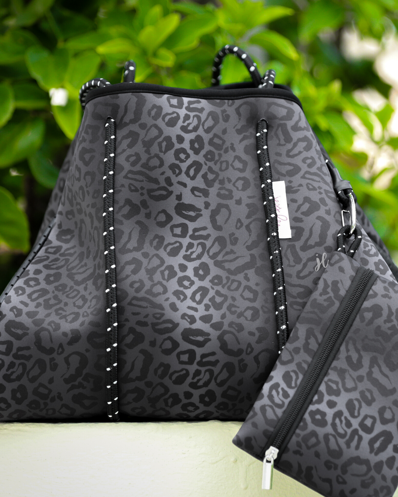 ultra-lightweight tote made of neoprene with a removable hard bottom liner, paracord shoulder straps, removable matching zippered pouch, magnetic side snap closures in silver hardware, and black interior (tote and pouch) on a black on black leopard print pattern