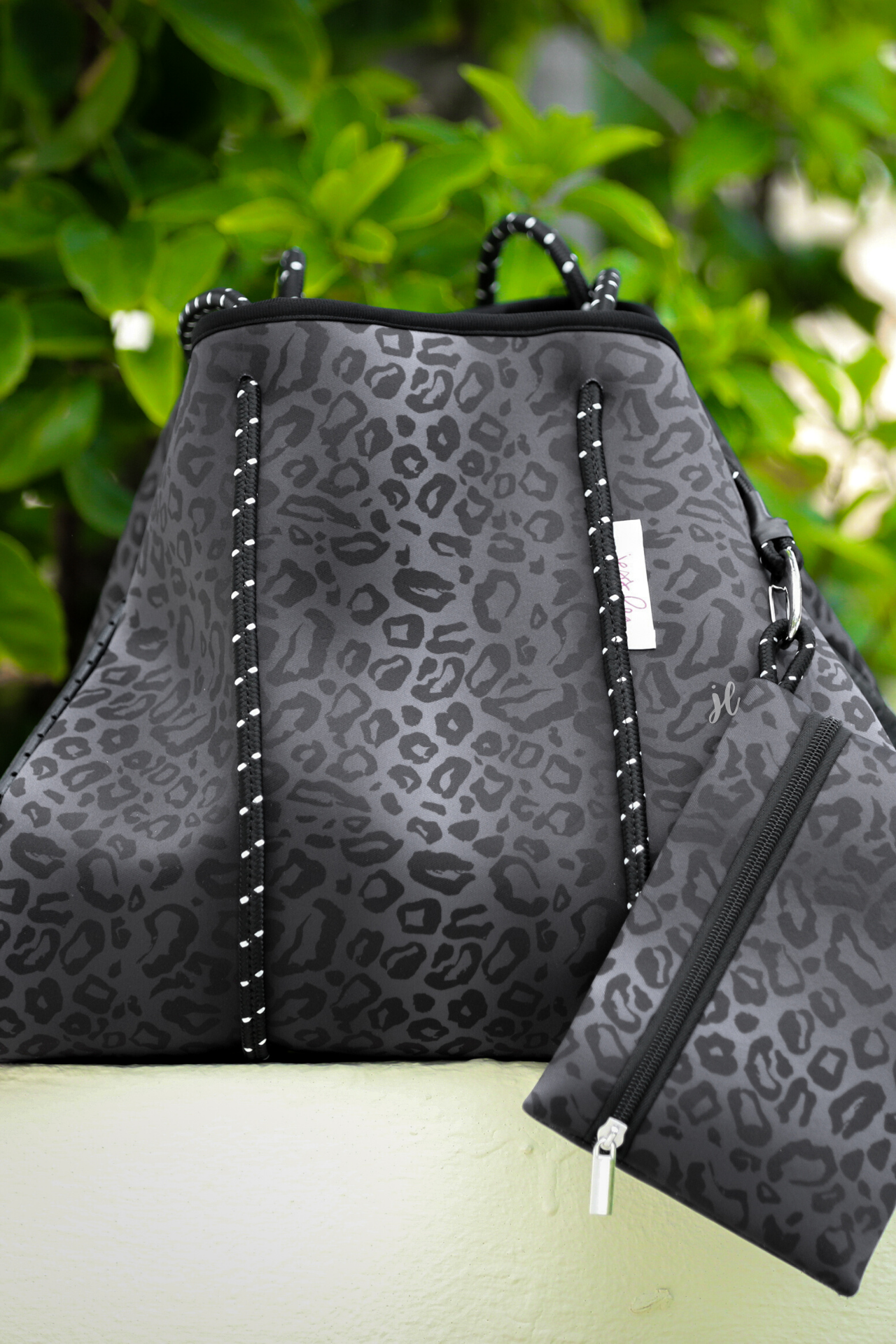 ultra-lightweight tote made of neoprene with a removable hard bottom liner, paracord shoulder straps, removable matching zippered pouch, magnetic side snap closures in silver hardware, and black interior (tote and pouch) on a black on black leopard print pattern