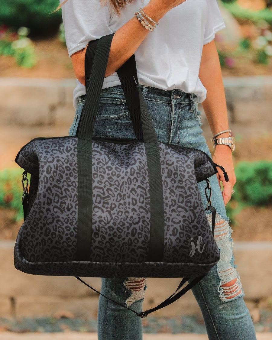 lightweight weekender bag made of neoprene with black shoulder straps, an adjustable black clip strap, two interior open side pockets, one interior zipper pocket, and zipper closure on a black on black leopard print fabric with black hardware