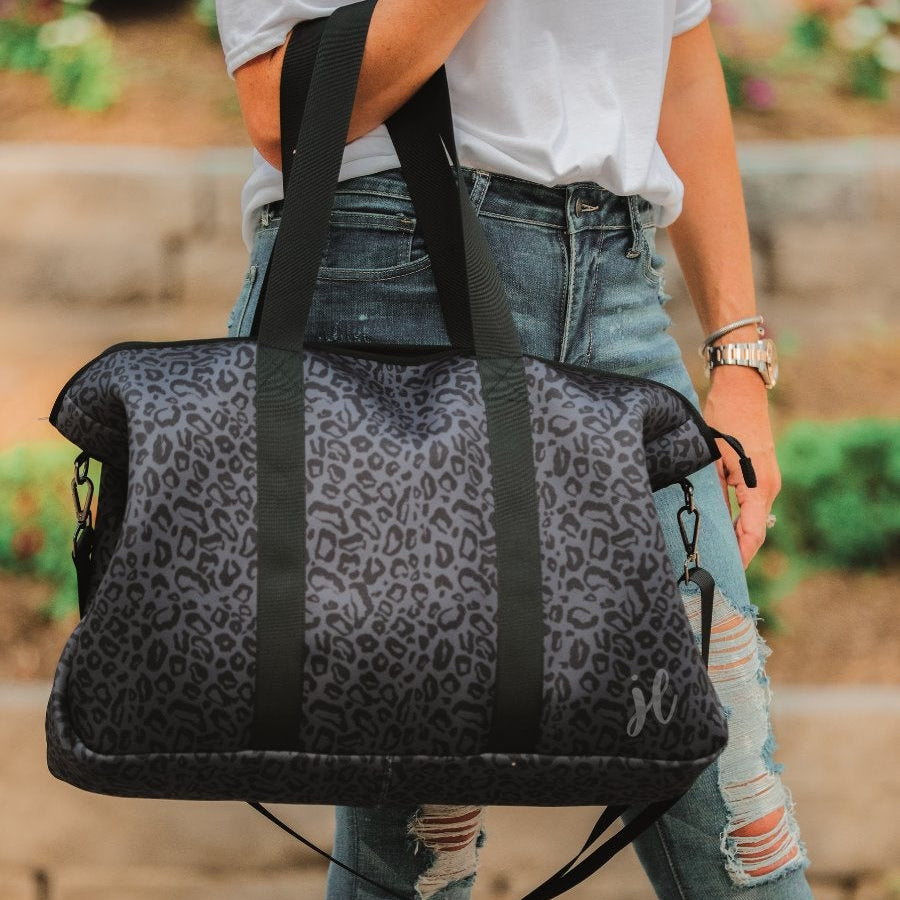 lightweight weekender bag made of neoprene with black shoulder straps, an adjustable black clip strap, two interior open side pockets, one interior zipper pocket, and zipper closure on a black on black leopard print fabric with black hardware