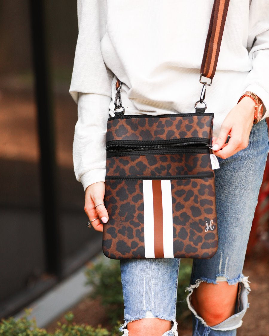 spacious crossbody bag in a black and brown leopard print made of neoprene with an adjustable strap (tonal brown), two front pockets (one zipper, one open), and racing stripes (brown and white) on a black and brown leopard print pattern