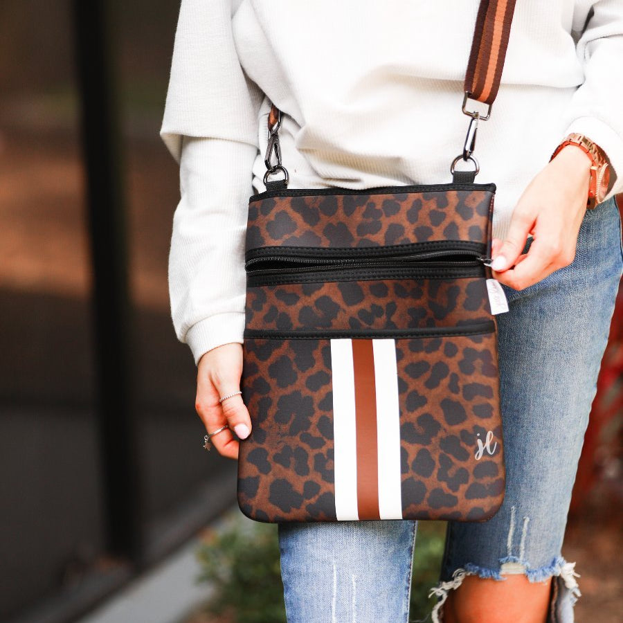 spacious crossbody bag in a black and brown leopard print made of neoprene with an adjustable strap (tonal brown), two front pockets (one zipper, one open), and racing stripes (brown and white) on a black and brown leopard print pattern