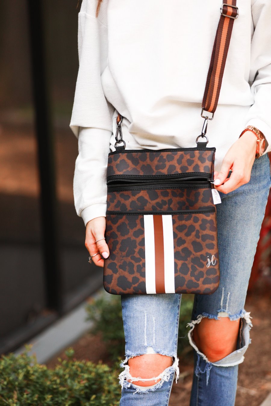 spacious crossbody bag in a black and brown leopard print made of neoprene with an adjustable strap (tonal brown), two front pockets (one zipper, one open), and racing stripes (brown and white) on a black and brown leopard print pattern