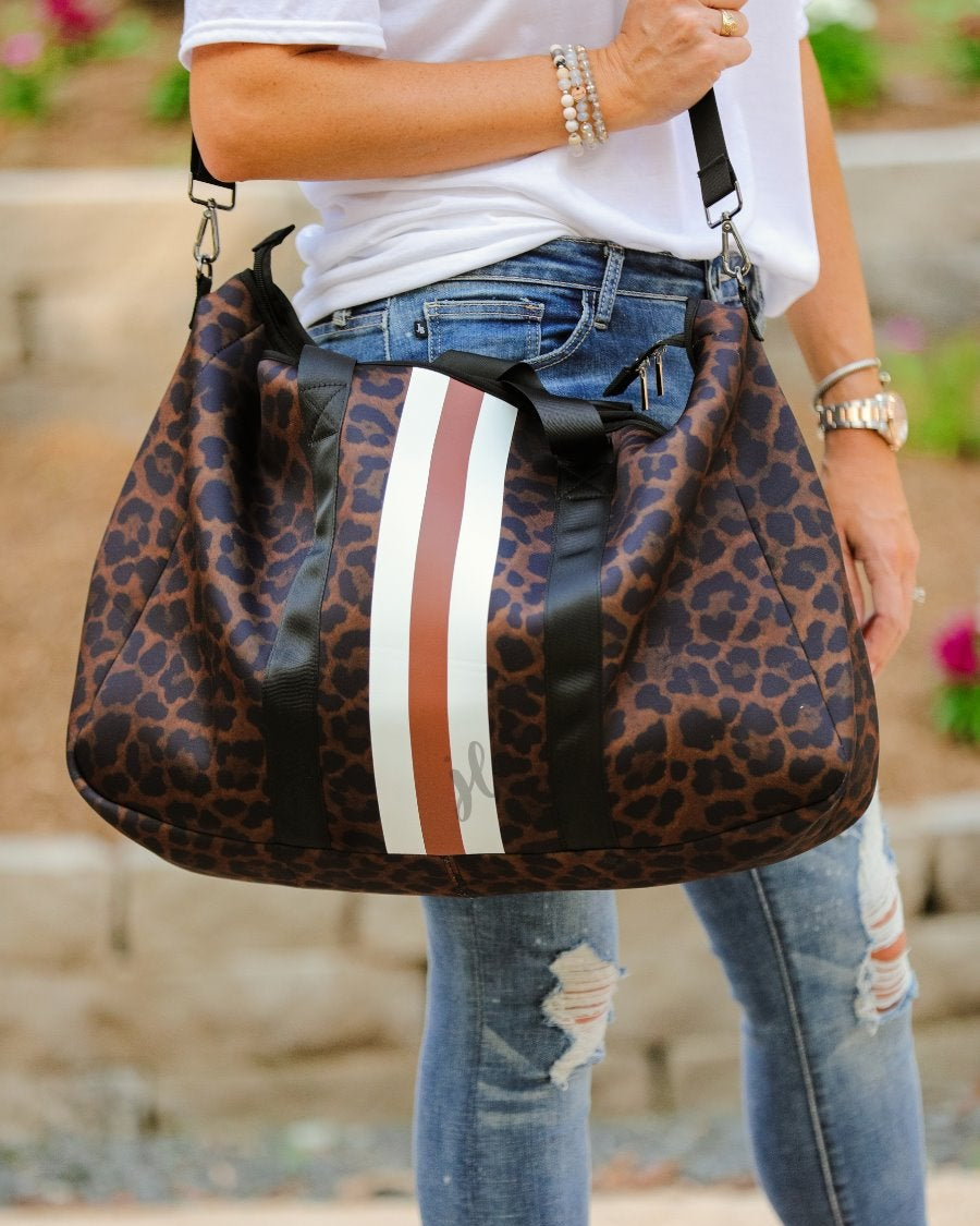 lightweight weekender bag made of neoprene with black shoulder straps, an adjustable black clip strap, two interior open side pockets, one interior zipper pocket, zipper closure, and racing stripes (brown and white) on a black on brown leopard print fabric with black hardware