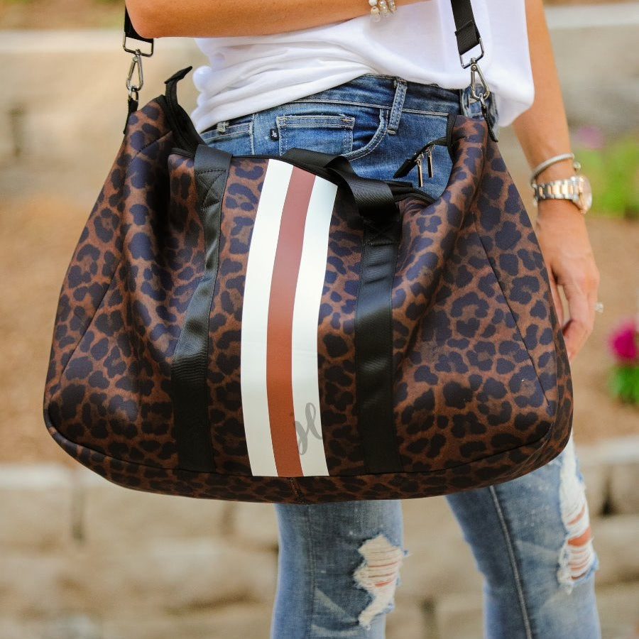 lightweight weekender bag made of neoprene with black shoulder straps, an adjustable black clip strap, two interior open side pockets, one interior zipper pocket, zipper closure, and racing stripes (brown and white) on a black on brown leopard print fabric with black hardware