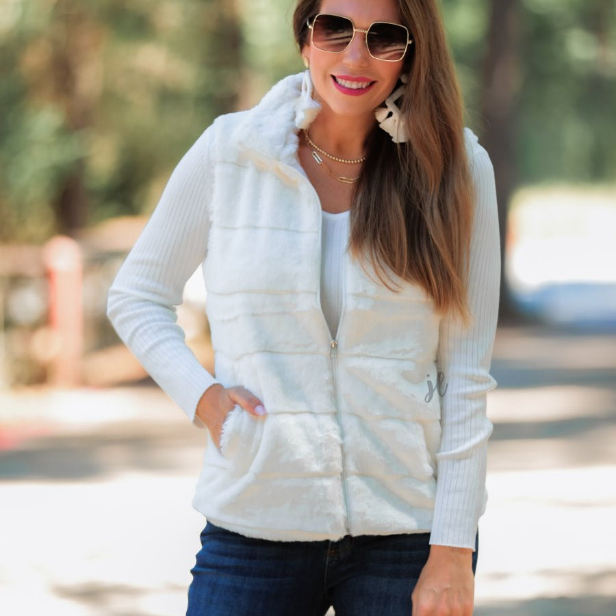 a sleeveless faux fur vest with a foldable collar, a front zip, sublte horizontal paneling, and front pockets made of super soft fabric that fits slightly oversized in white