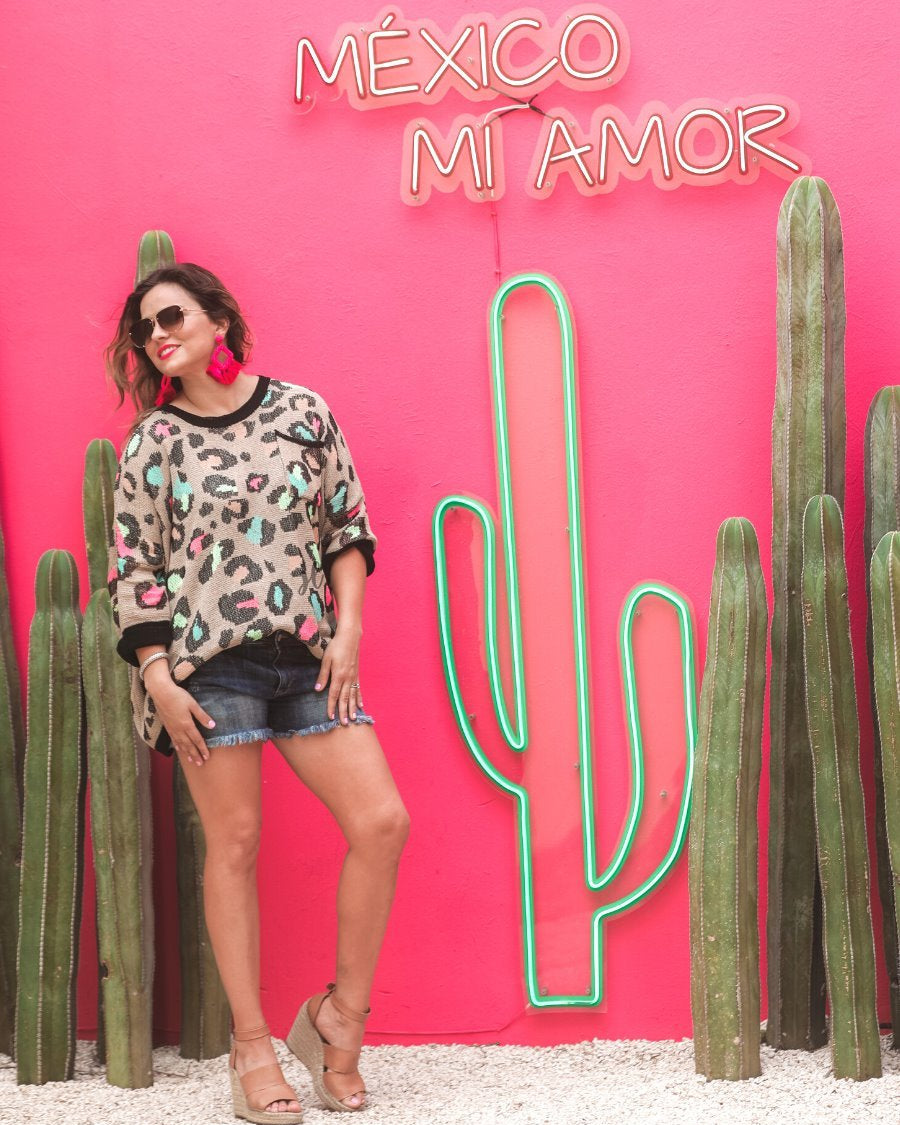 three quarter cuffed sleeve sweater made from unlined loose knit material with a rounded neckline, a chest pocket, and an oversized fit on a multicolor leopard pattern (blue, green, pink, and orange), a black accent color (chest pocket trim, cuffs, and hem), and a brown base