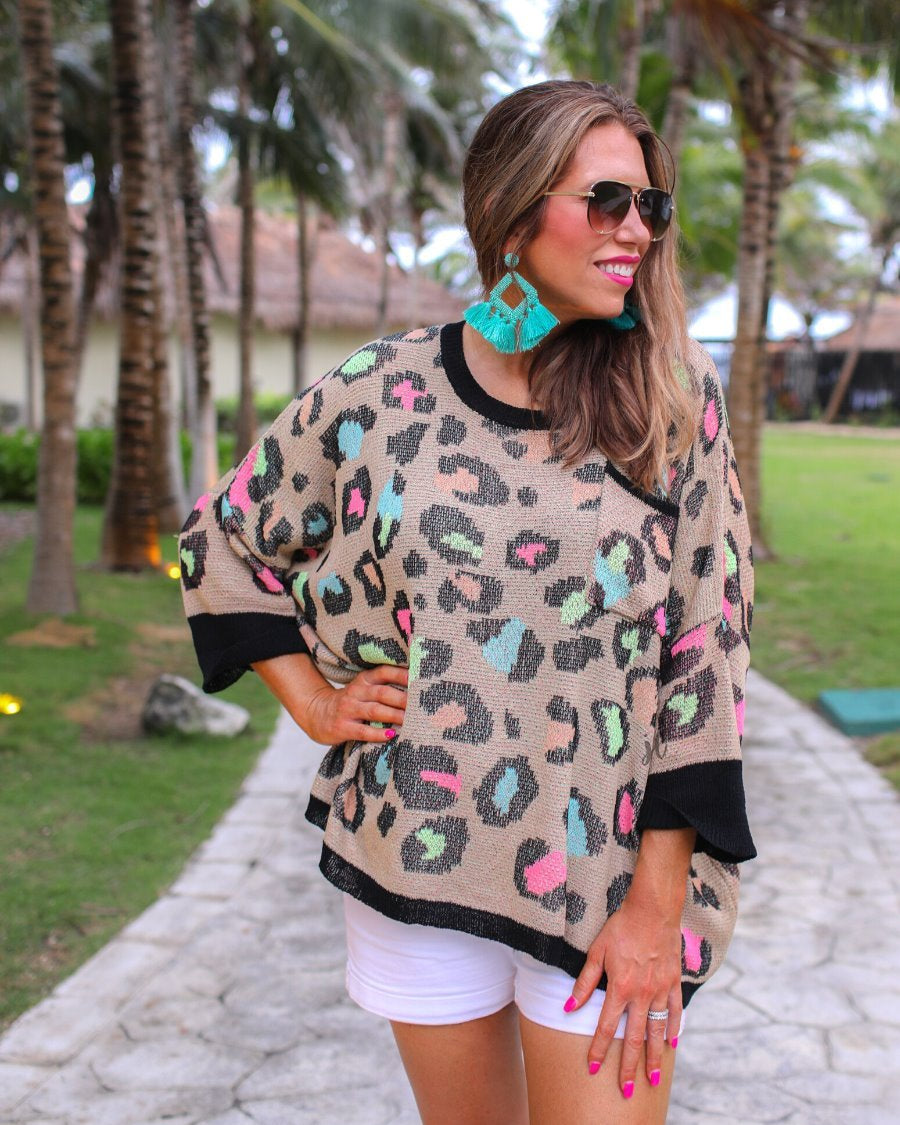 three quarter cuffed sleeve sweater made from unlined loose knit material with a rounded neckline, a chest pocket, and an oversized fit on a multicolor leopard pattern (blue, green, pink, and orange), a black accent color (chest pocket trim, cuffs, and hem), and a brown base