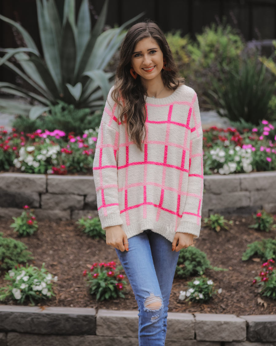 a plush knit long-sleeve sweater with a pink and ivory plaid pattern, relaxed drop-shoulder fit, classic round neckline, and ribbed detailing at the cuffs, sleeves, and hemline for a clean, polished finish
