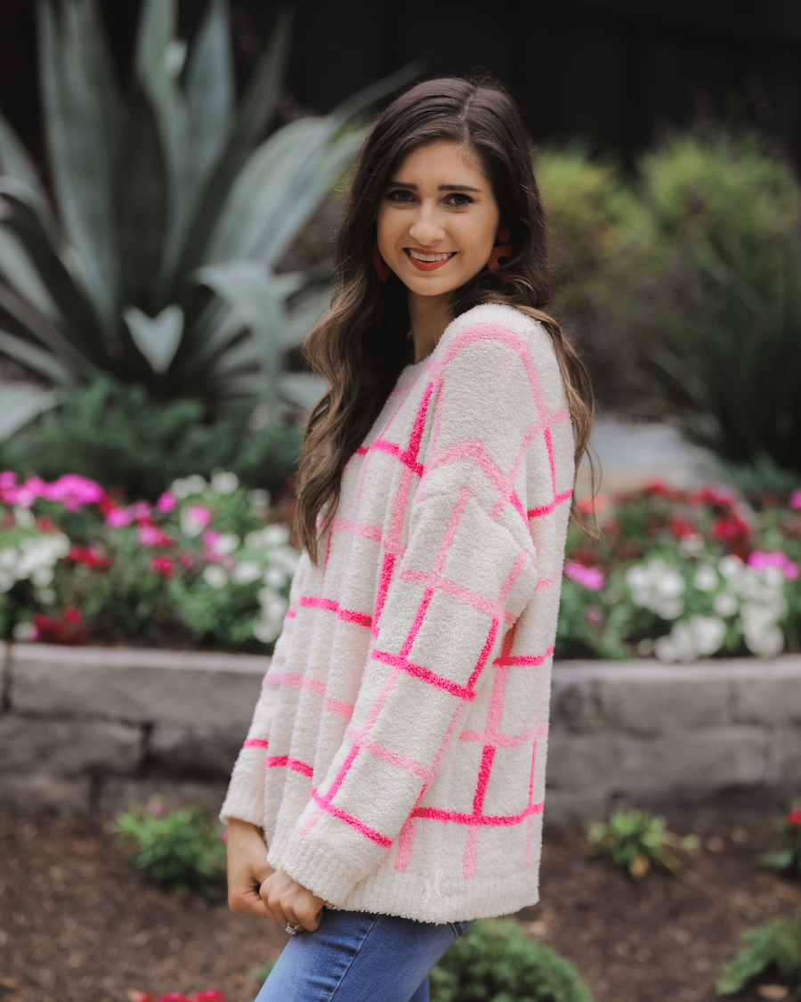 a plush knit long-sleeve sweater with a pink and ivory plaid pattern, relaxed drop-shoulder fit, classic round neckline, and ribbed detailing at the cuffs, sleeves, and hemline for a clean, polished finish