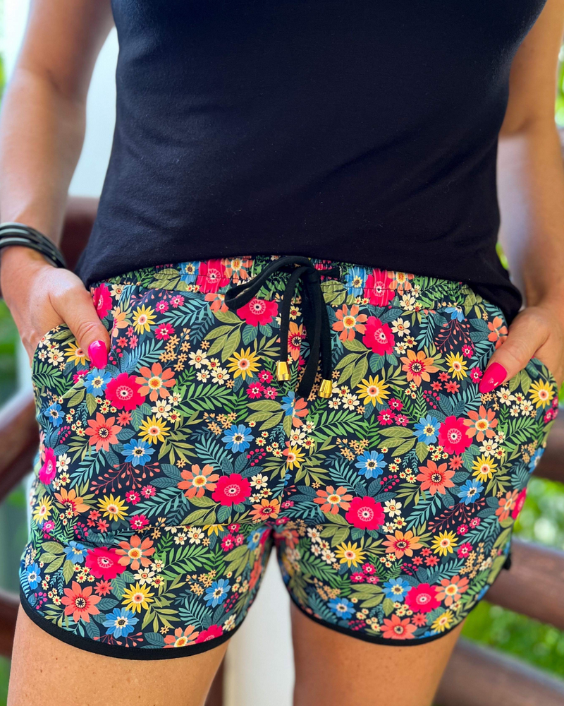 mid-rise relaxed fit activewear shorts with side pockets and a tie waist drawstring with gold bead accents, designed on a mix of multicolor floral and palm leaves pattern print fabric, navy base, and black trim