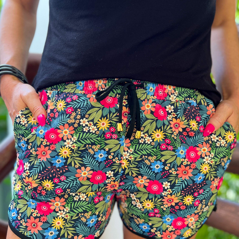 mid-rise relaxed fit activewear shorts with side pockets and a tie waist drawstring with gold bead accents, designed on a mix of multicolor floral and palm leaves pattern print fabric, navy base, and black trim