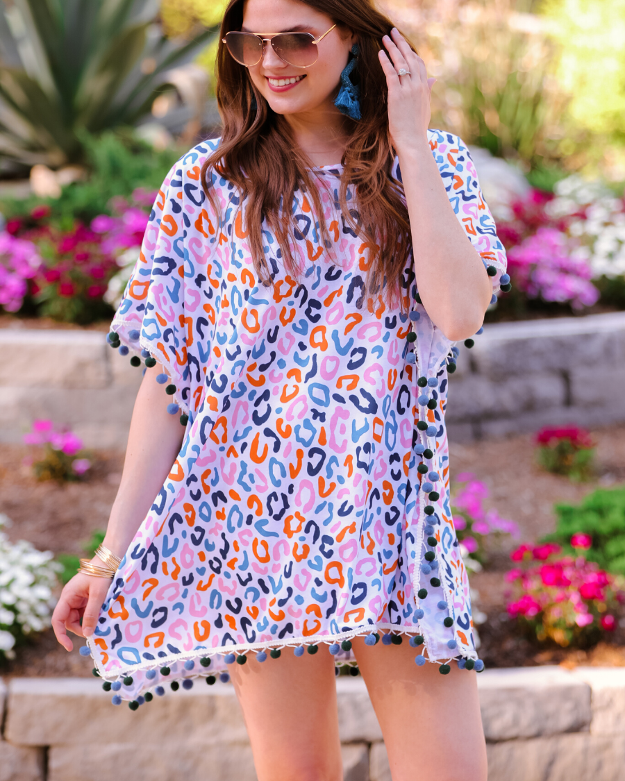 lightweight coverup made of breezy material with alternating dark blue and dark green pom pom decorative trim and multicolor leopard print pattern on a white background
