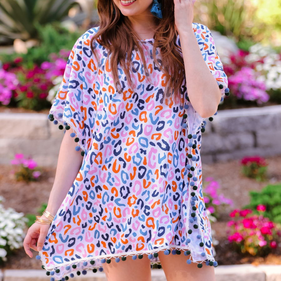 lightweight coverup made of breezy material with alternating dark blue and dark green pom pom decorative trim and multicolor leopard print pattern on a white background