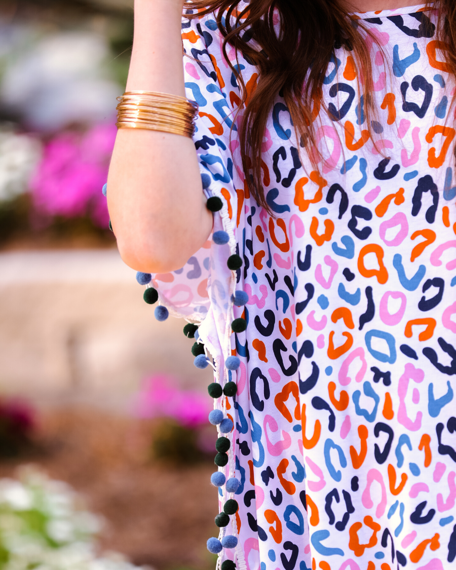 lightweight coverup made of breezy material with alternating dark blue and dark green pom pom decorative trim and multicolor leopard print pattern on a white background