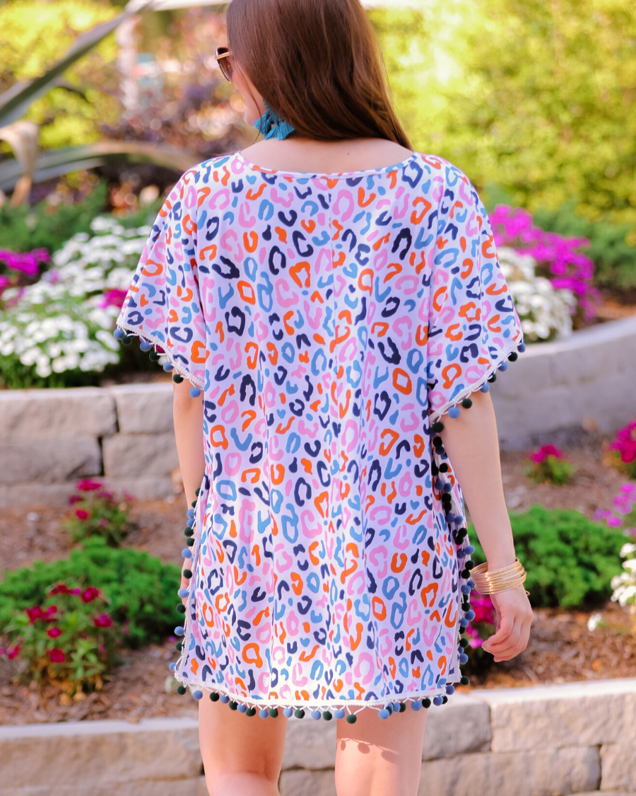 lightweight coverup made of breezy material with alternating dark blue and dark green pom pom decorative trim and multicolor leopard print pattern on a white background