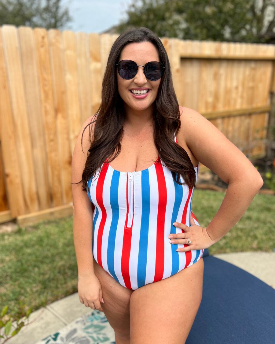 a white one-piece swimsuit made of stretchy material with thick straps, a scoop neckline, a zip front, alternating red and blue vertical stripes, and removable padding