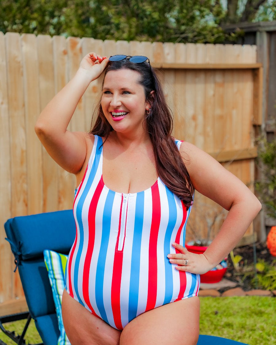 a white one-piece swimsuit made of stretchy material with thick straps, a scoop neckline, a zip front, alternating red and blue vertical stripes, and removable padding