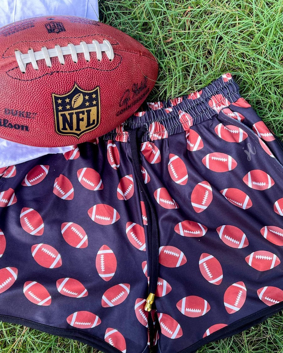 mid-rise relaxed fit activewear shorts with side pockets and a tie waist drawstring with gold bead accent, designed with a football pattern print, black base, and black trim