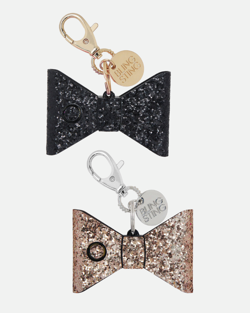 small compact personal safety alarm in a glittery bow shape with a keychain clip, an oversized lobster clip, a pink trigger button, and an LED safety light button operating on a 12-volt A23 battery available in black or rose gold