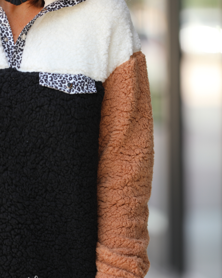 a long-sleeve fluffy pullover with a quarter-leopard snap closure, a faux leopard front chest pocket, a foldable collar, and convenient side pockets on a black, taupe, and white color block fabric that is slightly oversized