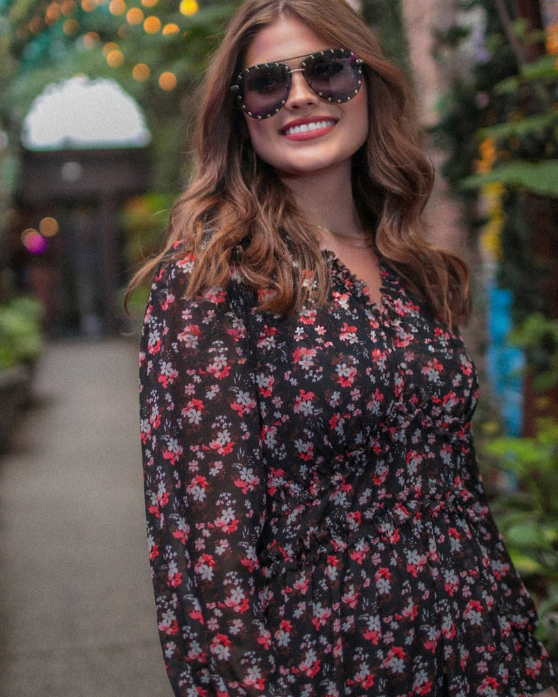 a black floral pattern print dress with sheer long sleeves, smocked cuffs, a ruffle v-neckline, back zipper, tiered ruffle hemline, and full lining with no stretch