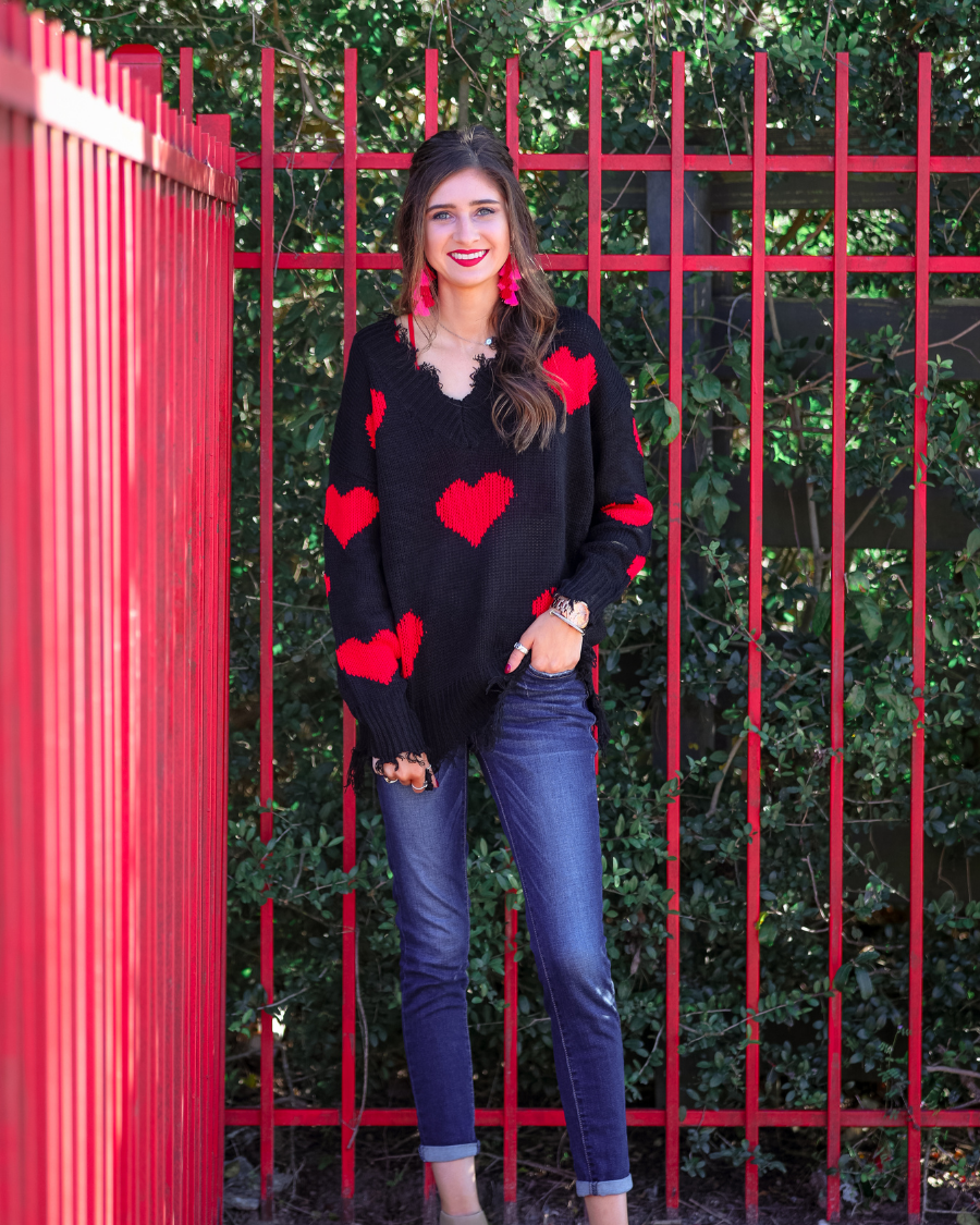 a long-sleeve sweater crafted from ultra soft, stretchy knit fabric with striking red heart motifs across a rich black knit, a relaxed drop-shoulder fit, and distressed ribbed detailing on v-neckline, cuffs, and hemline