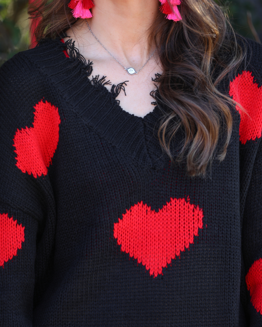 a long-sleeve sweater crafted from ultra soft, stretchy knit fabric with striking red heart motifs across a rich black knit, a relaxed drop-shoulder fit, and distressed ribbed detailing on v-neckline, cuffs, and hemline
