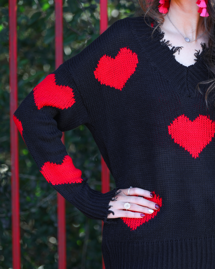 a long-sleeve sweater crafted from ultra soft, stretchy knit fabric with striking red heart motifs across a rich black knit, a relaxed drop-shoulder fit, and distressed ribbed detailing on v-neckline, cuffs, and hemline