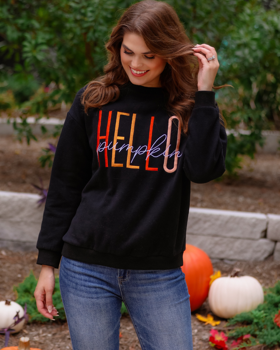 black long-sleeve sweatshirt made of soft mid-weight fabric with a crew neckline, straight hem, and colorful embroidered mix of 'HELLO' in uppercase letters (orange, rose gold, and yellow) and 'PUMPKIN' in white script handwriting