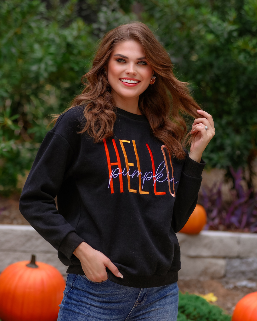 black long-sleeve sweatshirt made of soft mid-weight fabric with a crew neckline, straight hem, and colorful embroidered mix of 'HELLO' in uppercase letters (orange, rose gold, and yellow) and 'PUMPKIN' in white script handwriting