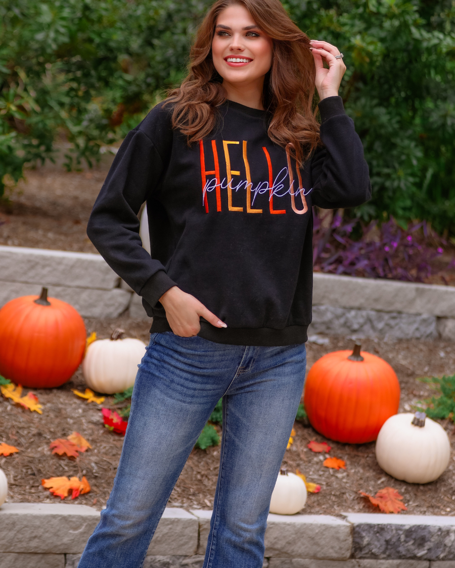 black long-sleeve sweatshirt made of soft mid-weight fabric with a crew neckline, straight hem, and colorful embroidered mix of 'HELLO' in uppercase letters (orange, rose gold, and yellow) and 'PUMPKIN' in white script handwriting