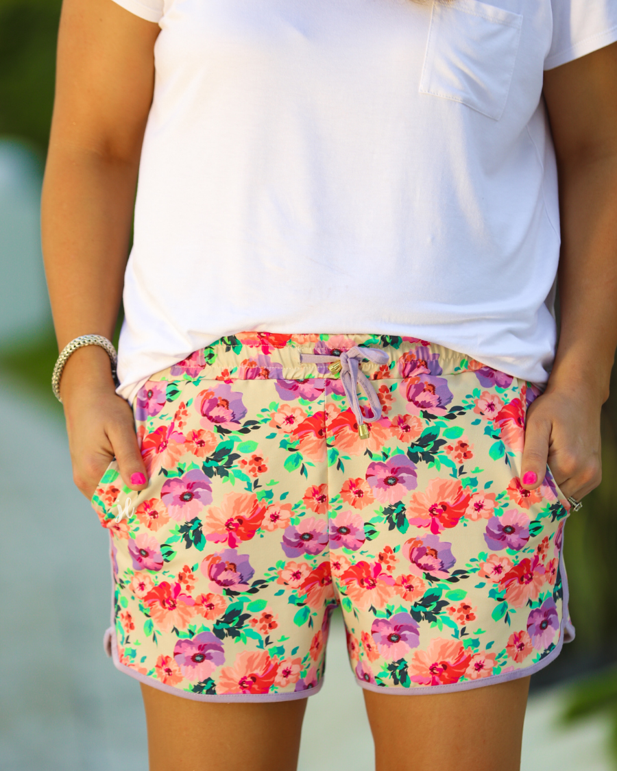 mid-rise relaxed fit activewear shorts with side pockets and a tie waist drawstring with gold bead accents, designed on a pastel floral pattern print fabric, cream base, and lavender trim