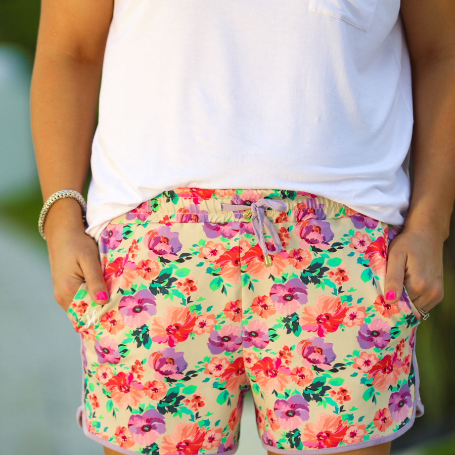 mid-rise relaxed fit activewear shorts with side pockets and a tie waist drawstring with gold bead accents, designed on a pastel floral pattern print fabric, cream base, and lavender trim