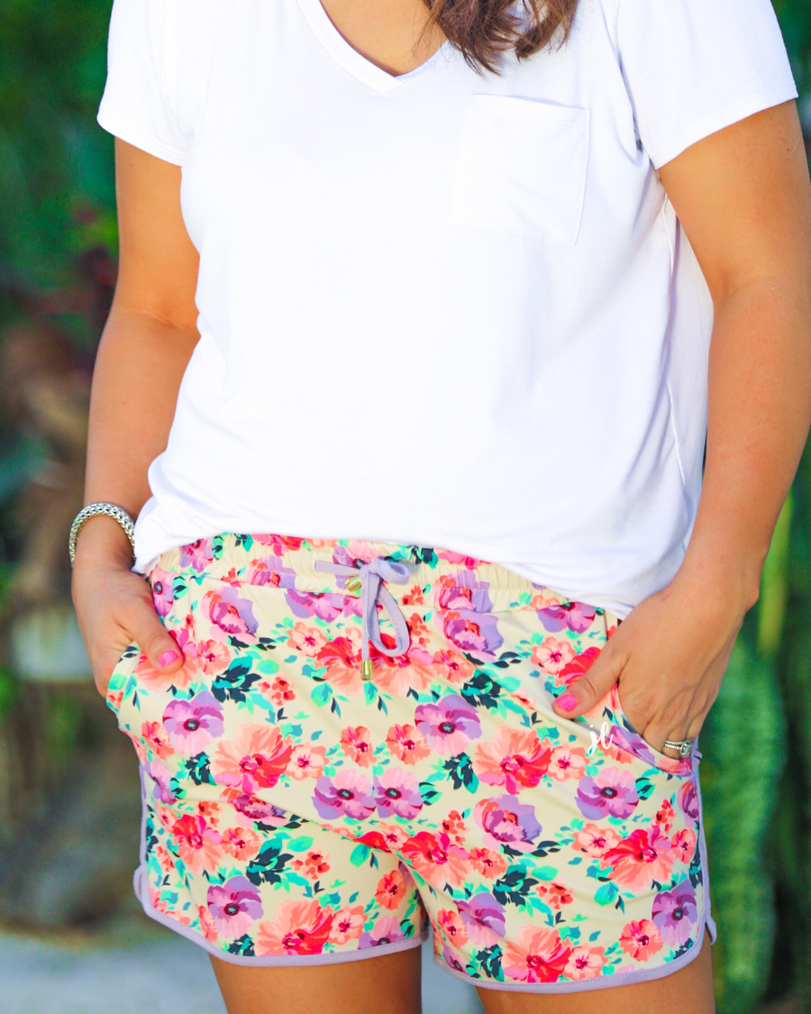 mid-rise relaxed fit activewear shorts with side pockets and a tie waist drawstring with gold bead accents, designed on a pastel floral pattern print fabric, cream base, and lavender trim