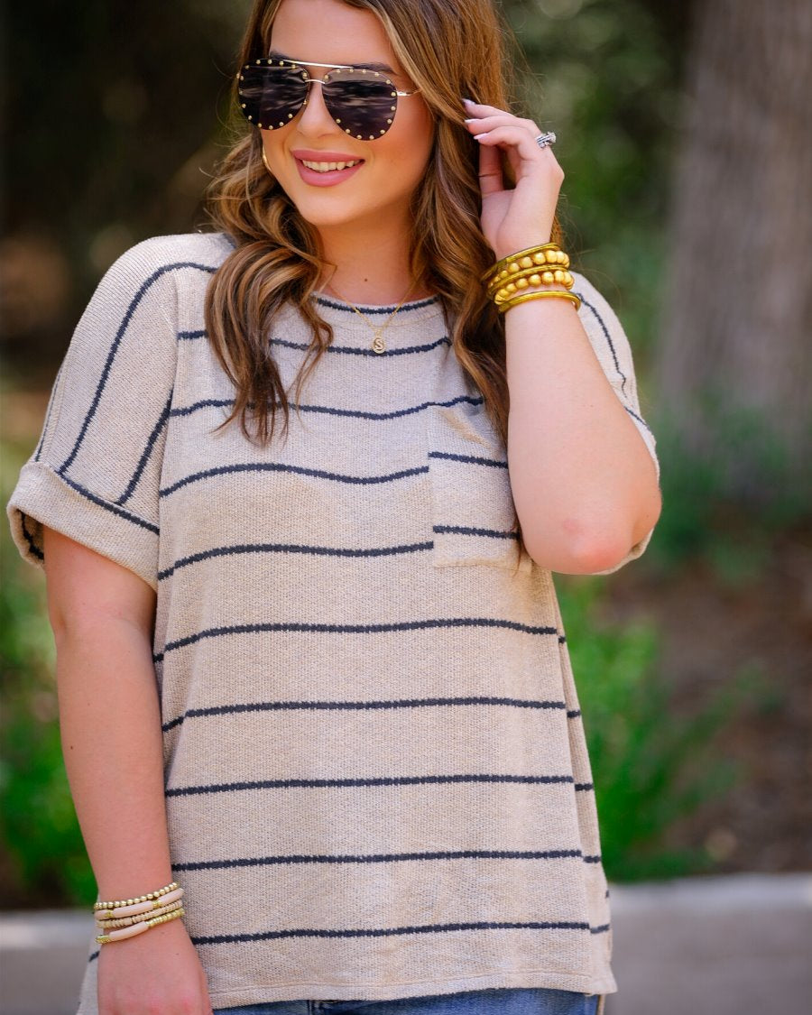 short-sleeve top made of a soft knit fabric with horizontal stripes, a rounded neckline, rolled cuffs, a front check pocket, and a relaxed silhouette on a taupe base