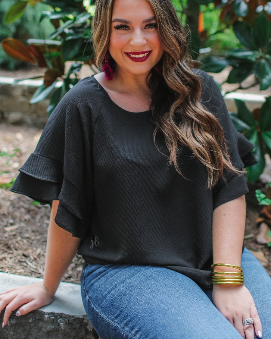 a black true-to-size blouse crafted from lightweight textured fabric with a rounded neckline, tiered ruffle sleeves, and a relaxed drape