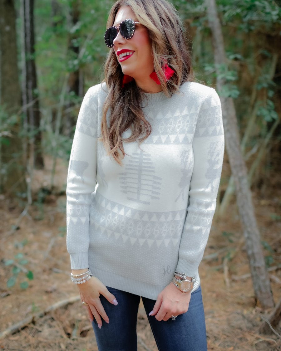 a grey long-sleeve heavy sweater with a ribbed detailing on the neckline, cuffs, and hem on a festive grey and white holiday pattern fabric