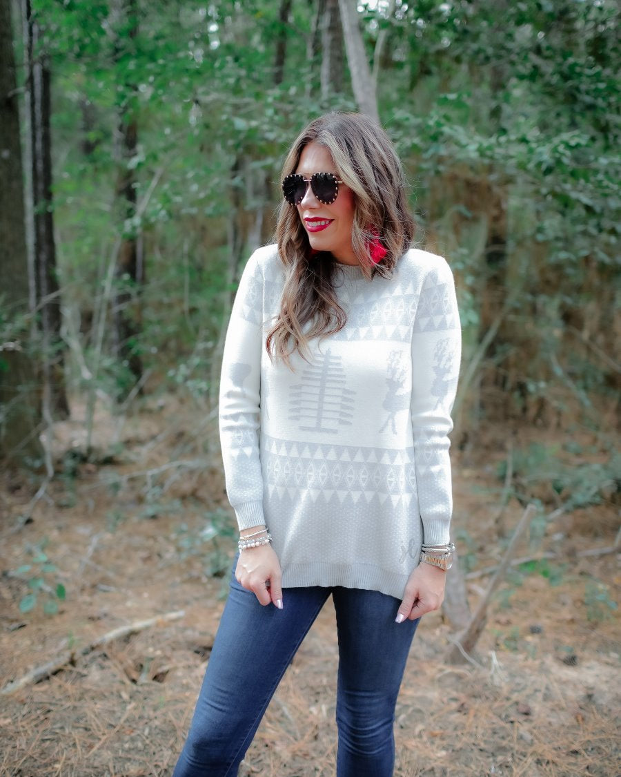a grey long-sleeve heavy sweater with a ribbed detailing on the neckline, cuffs, and hem on a festive grey and white holiday pattern fabric
