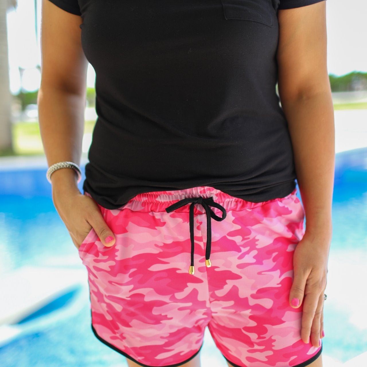 mid-rise relaxed fit activewear shorts with side pockets and a tie waist drawstring with gold bead accents, designed on a pink camo pattern print fabric, pink base, and black trim