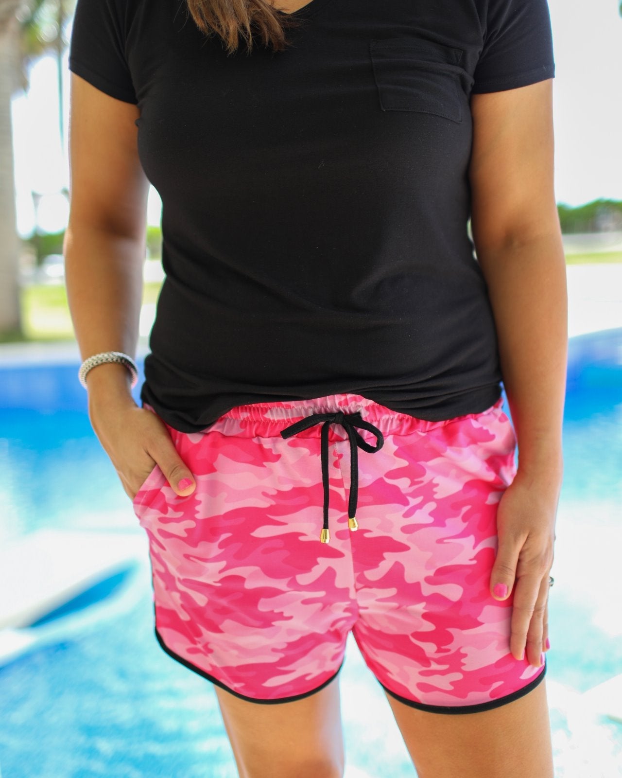 mid-rise relaxed fit activewear shorts with side pockets and a tie waist drawstring with gold bead accents, designed on a pink camo pattern print fabric, pink base, and black trim