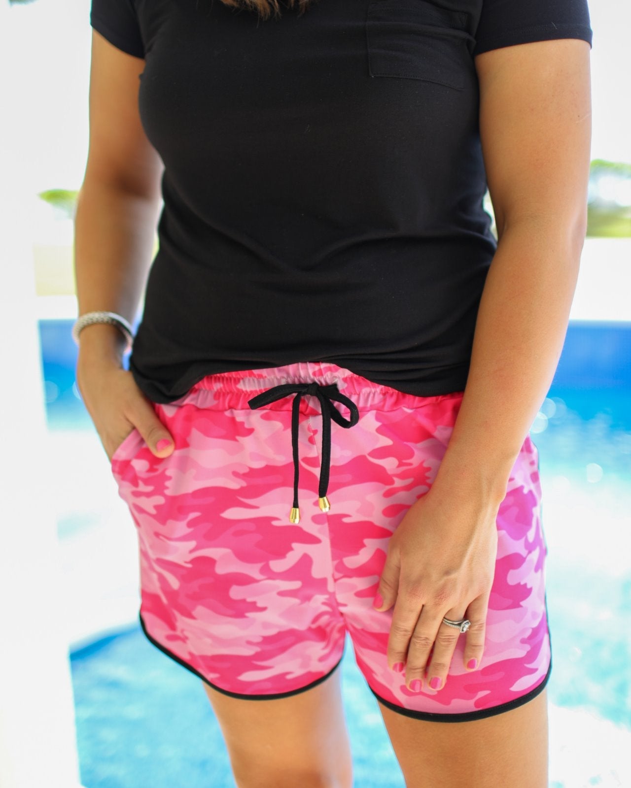mid-rise relaxed fit activewear shorts with side pockets and a tie waist drawstring with gold bead accents, designed on a pink camo pattern print fabric, pink base, and black trim