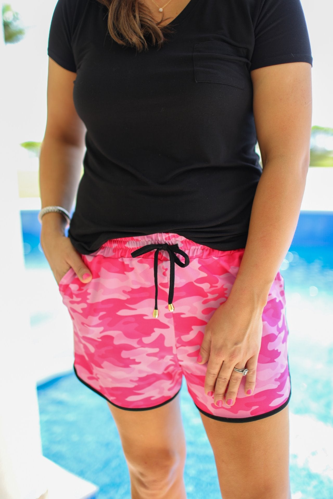 mid-rise relaxed fit activewear shorts with side pockets and a tie waist drawstring with gold bead accents, designed on a pink camo pattern print fabric, pink base, and black trim