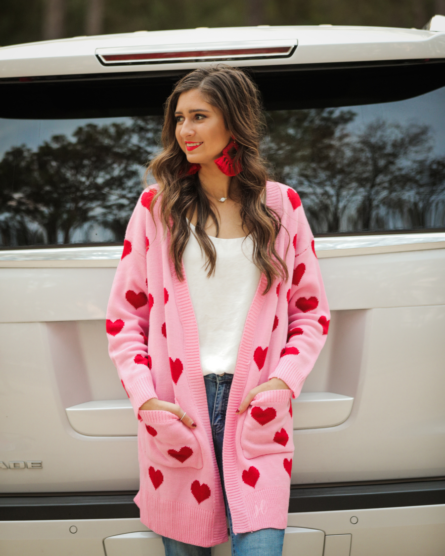 a long-sleeve open-front cardigan crafted on a soft pink knit fabric with a red heart pattern, functional front pockets, and ribbed detailing on the neckline, cuffs, and hemline