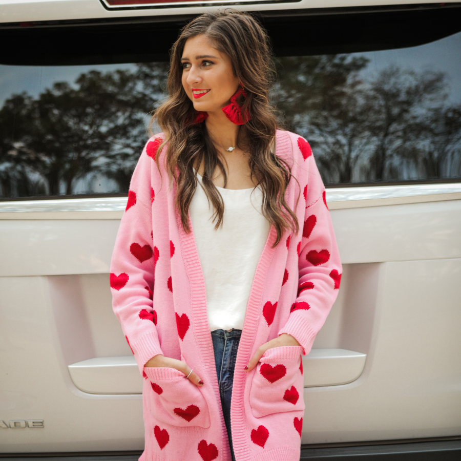 a long-sleeve open-front cardigan crafted on a soft pink knit fabric with a red heart pattern, functional front pockets, and ribbed detailing on the neckline, cuffs, and hemline