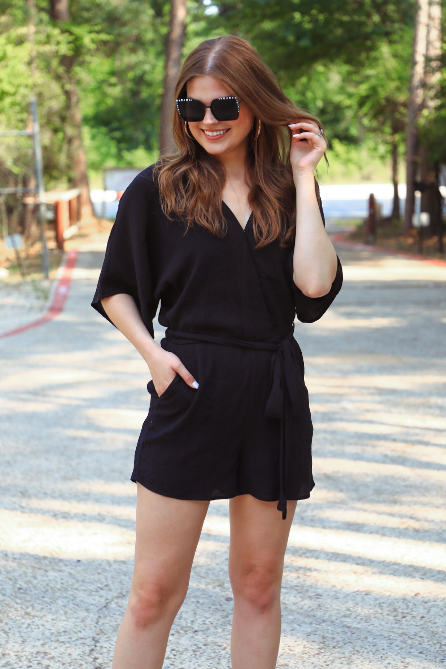 a black lined romper made of soft lightweight fabric with a surplice V-neckline, snap closure, relaxed flowy sleeves, waist-defining accent tie, side pockets, and a flattering easy-to-wear silhouette