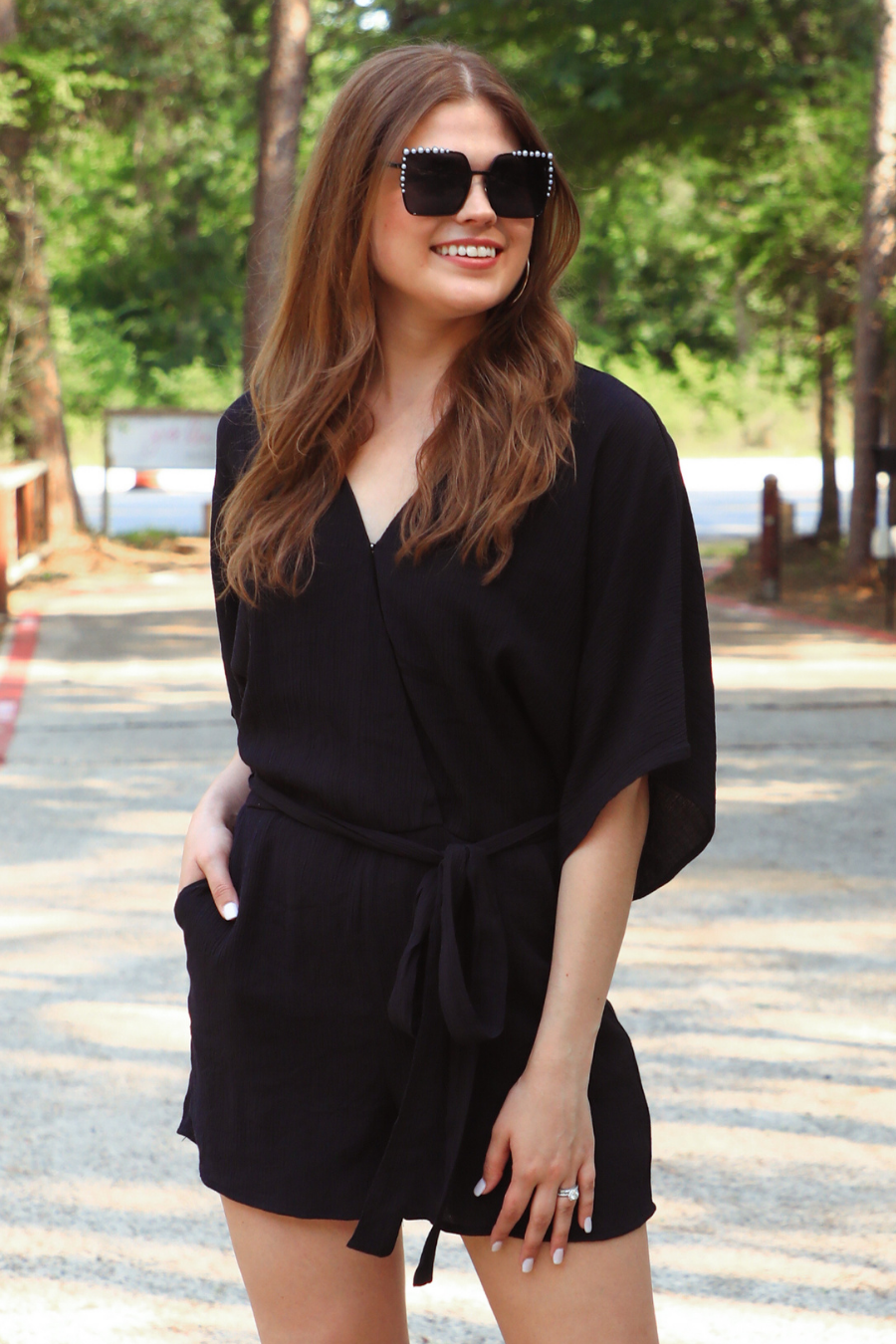 a black lined romper made of soft lightweight fabric with a surplice V-neckline, snap closure, relaxed flowy sleeves, waist-defining accent tie, side pockets, and a flattering easy-to-wear silhouette