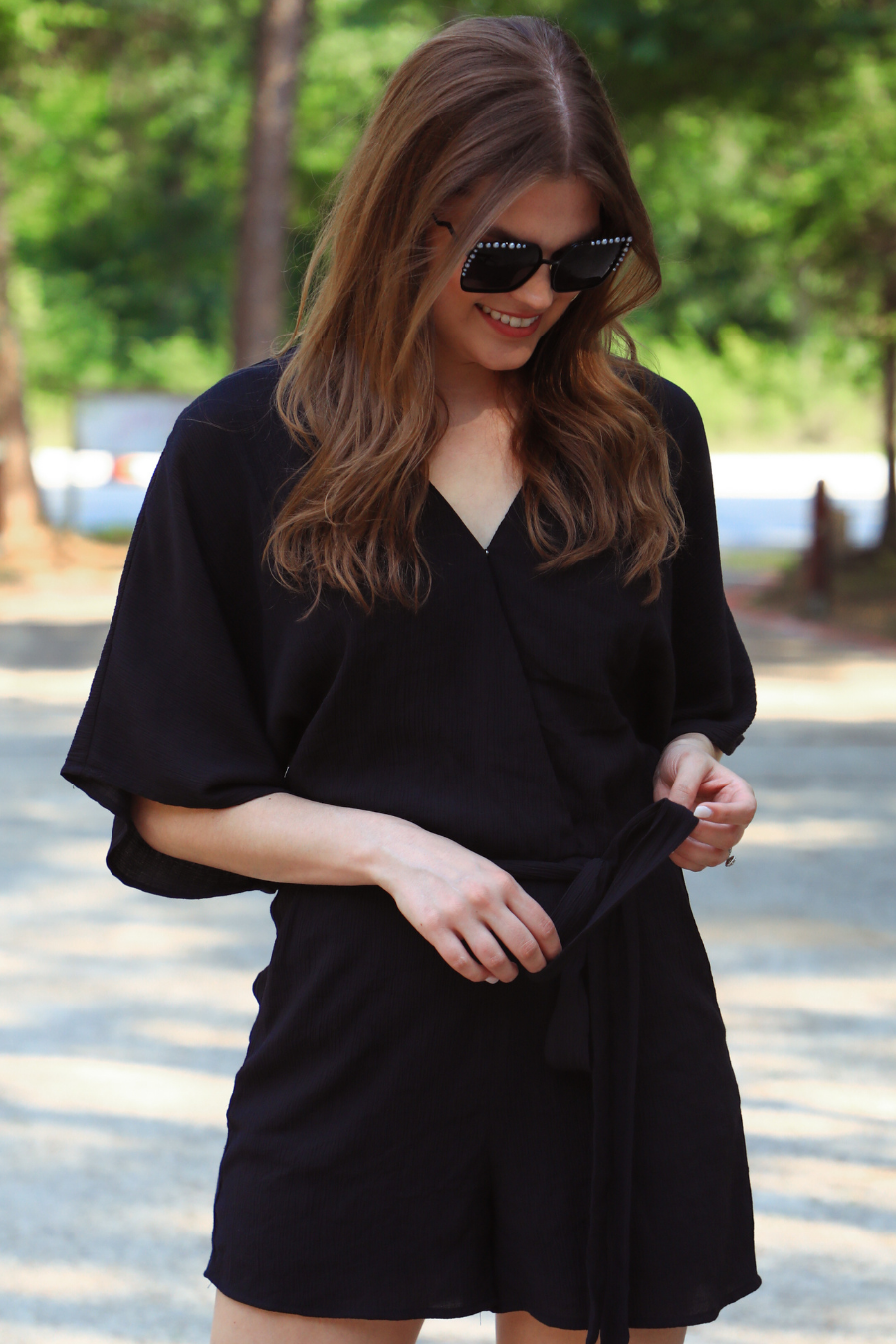 a black lined romper made of soft lightweight fabric with a surplice V-neckline, snap closure, relaxed flowy sleeves, waist-defining accent tie, side pockets, and a flattering easy-to-wear silhouette
