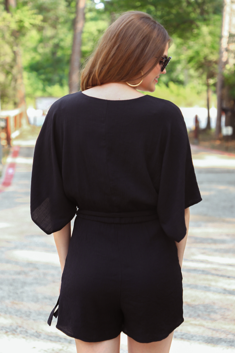 a black lined romper made of soft lightweight fabric with a surplice V-neckline, snap closure, relaxed flowy sleeves, waist-defining accent tie, side pockets, and a flattering easy-to-wear silhouette